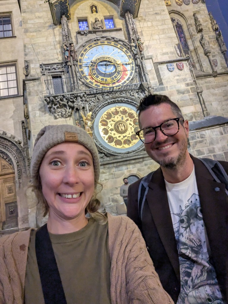 A smiling woman and man posing for a selfie in front of a historic clock tower at night.