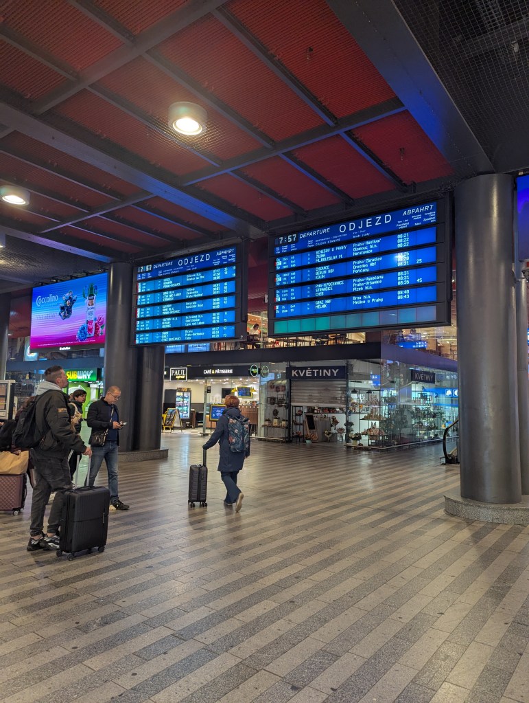 Interior view of a train station showing departure screens displaying train schedules and several travelers with luggage in the foreground.