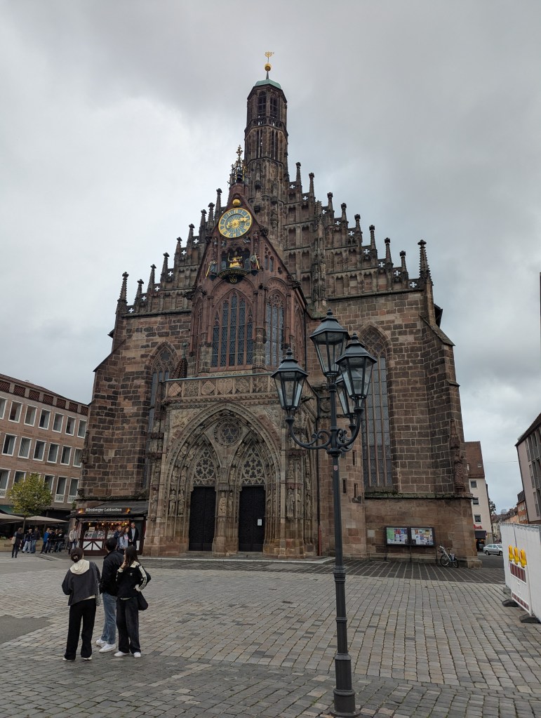 View of a historic church with intricate architecture in a cobblestone square, featuring a tall tower with a clock and spires, with a few people standing nearby.