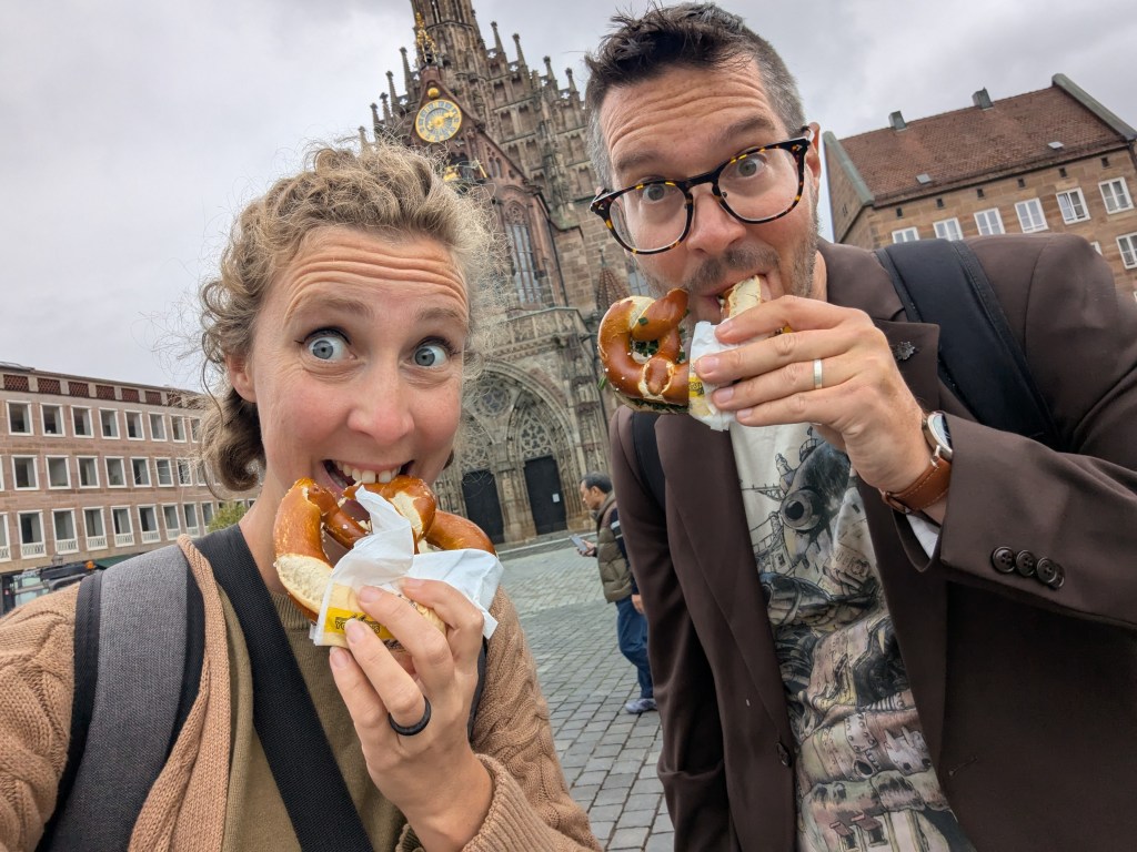 A couple happily posing with pretzels in front of a historic building in a city square on a cloudy day.