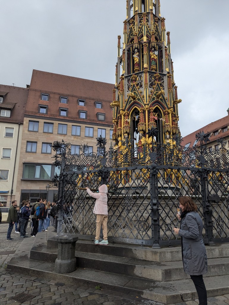 A child in a light pink jacket climbing on a decorative metal fence surrounding a tall, ornate monument with gold and red details, while people gather in the background.