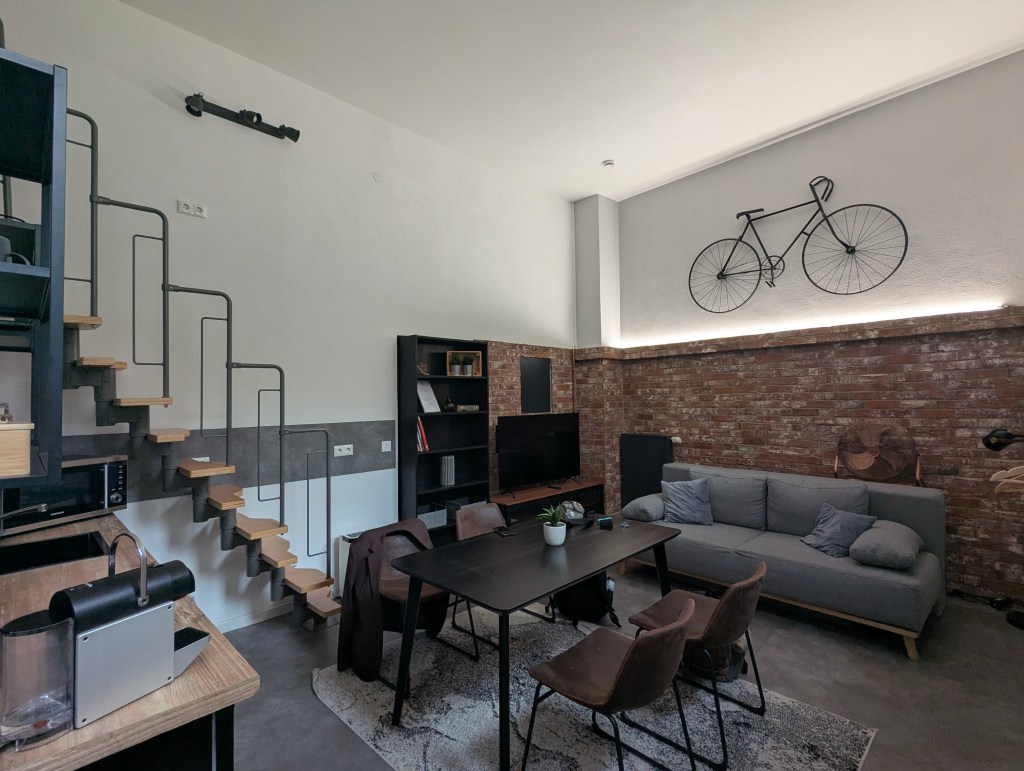 A modern living space featuring a staircase, a cozy gray sofa, a wooden dining table with chairs, and a decorative bicycle mounted on the wall.