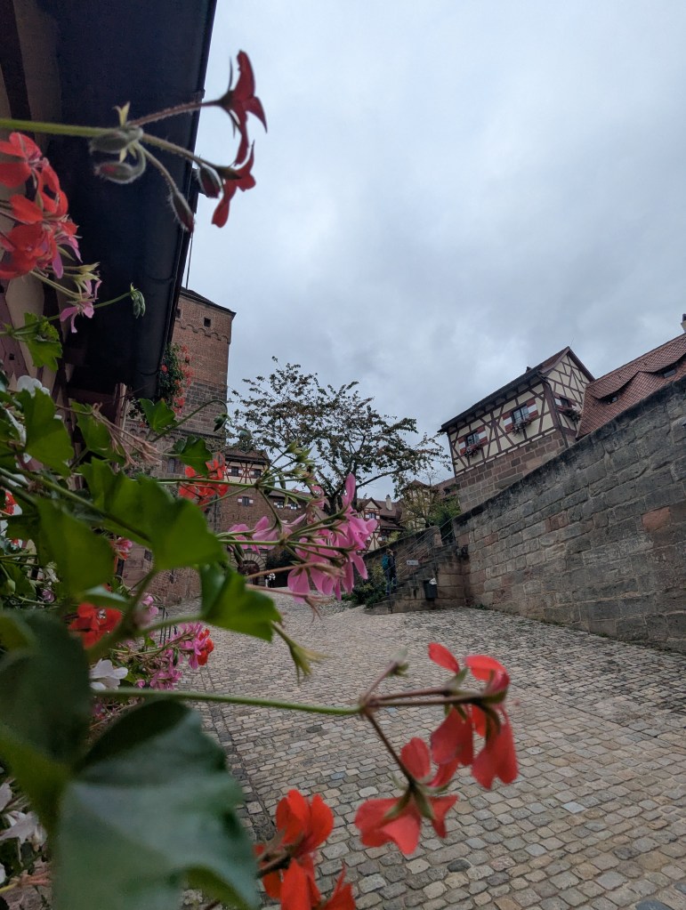 A cobblestone pathway lined with colorful flowers, leading towards historic buildings under a cloudy sky.