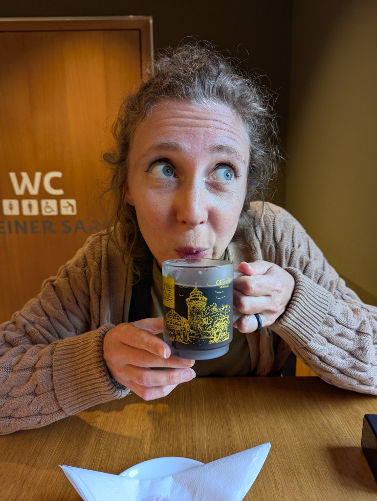 A woman with curly hair and blue eyes sipping from a decorative mug while sitting at a wooden table. The background features a sign indicating restrooms.