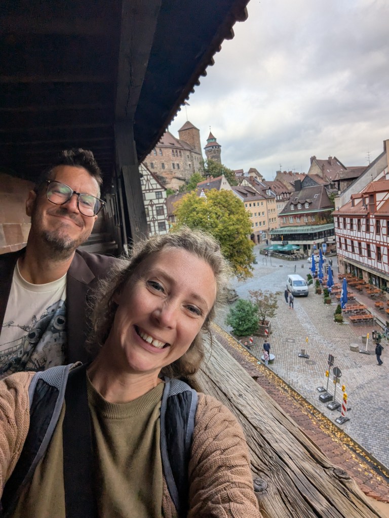 A smiling couple poses for a selfie on a wooden balcony overlooking a quaint street with historic buildings and a tower in the background, under a cloudy sky.