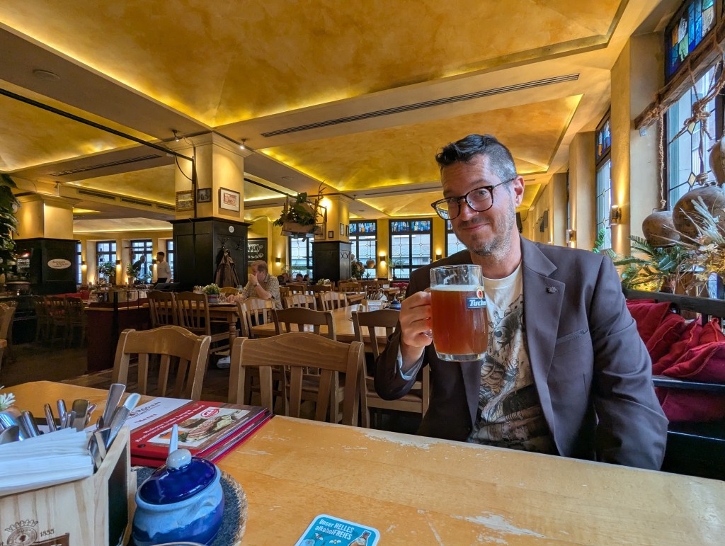 A man sitting at a wooden table in a warm and inviting restaurant, holding up a large beer mug. The interior features yellow lighting, wooden chairs, and decorative plants in the background.