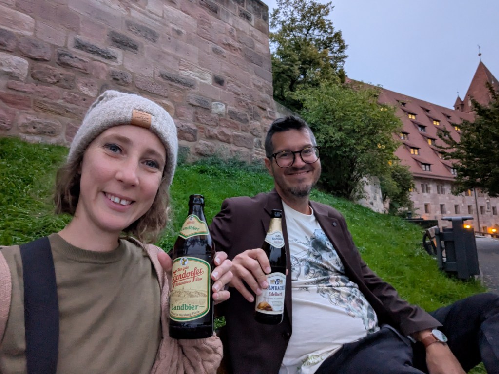A woman and a man sitting on grass near a stone wall, both smiling and holding bottles of beer.