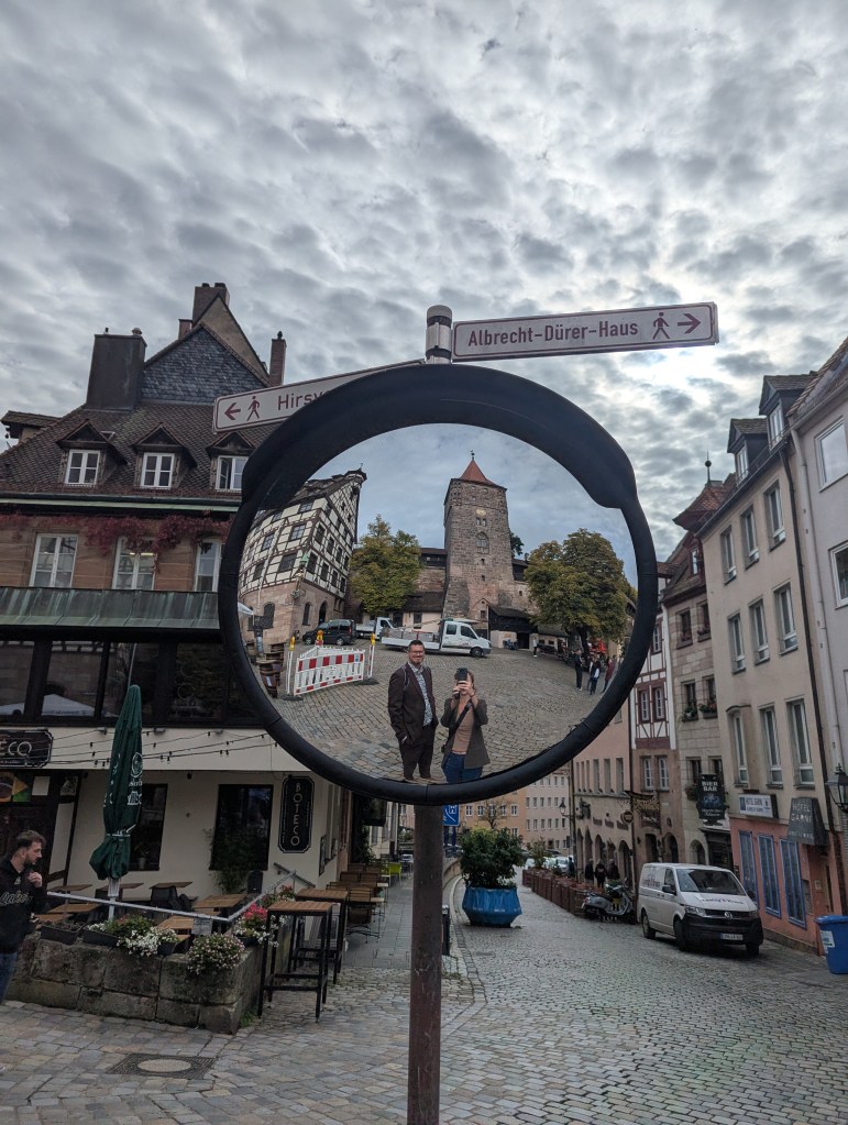 Reflected image in a convex mirror showing two people standing in a cobblestone street near a restaurant, with a historic tower and street signs in the background under a cloudy sky.
