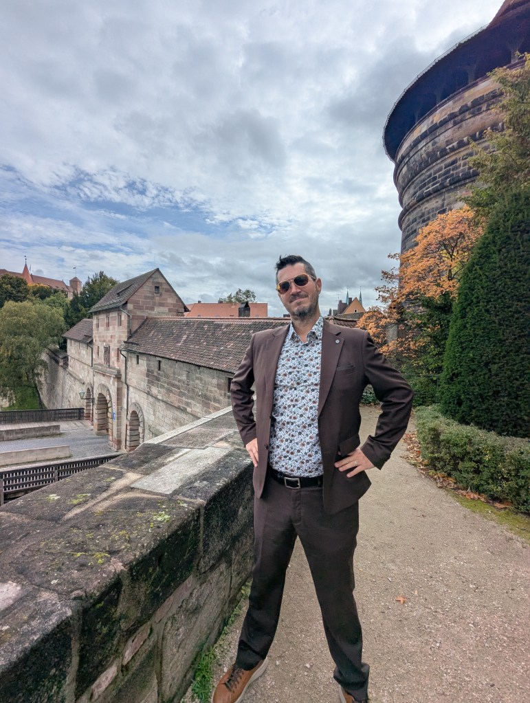 A man in stylish attire stands confidently near a historic stone wall with greenery and an old castle tower in the background under a cloudy sky.