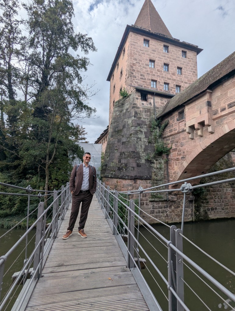 A person standing on a wooden bridge in front of a historic stone tower surrounded by greenery.