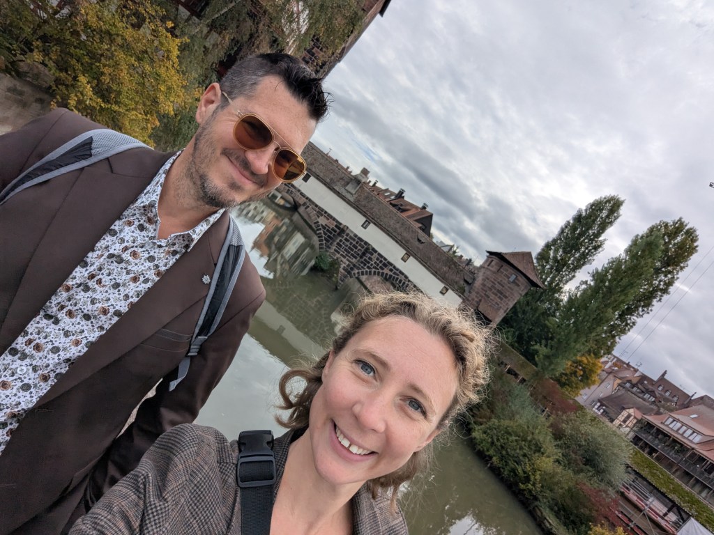 A smiling man in sunglasses and a patterned shirt stands beside a woman with curly hair, both photographed in front of a river and trees, with historic buildings in the background under a cloudy sky.
