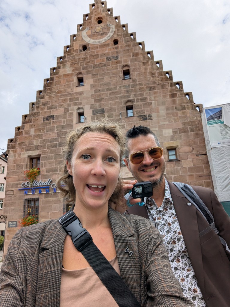 A cheerful couple taking a selfie in front of a historic building with a distinct architectural design in a picturesque setting.