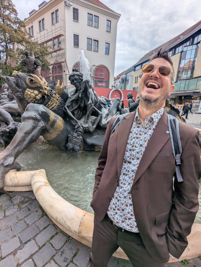A man in a brown suit and sunglasses stands laughing next to a large fountain featuring intricate sculptures, including a dragon and other figures, in an urban setting.