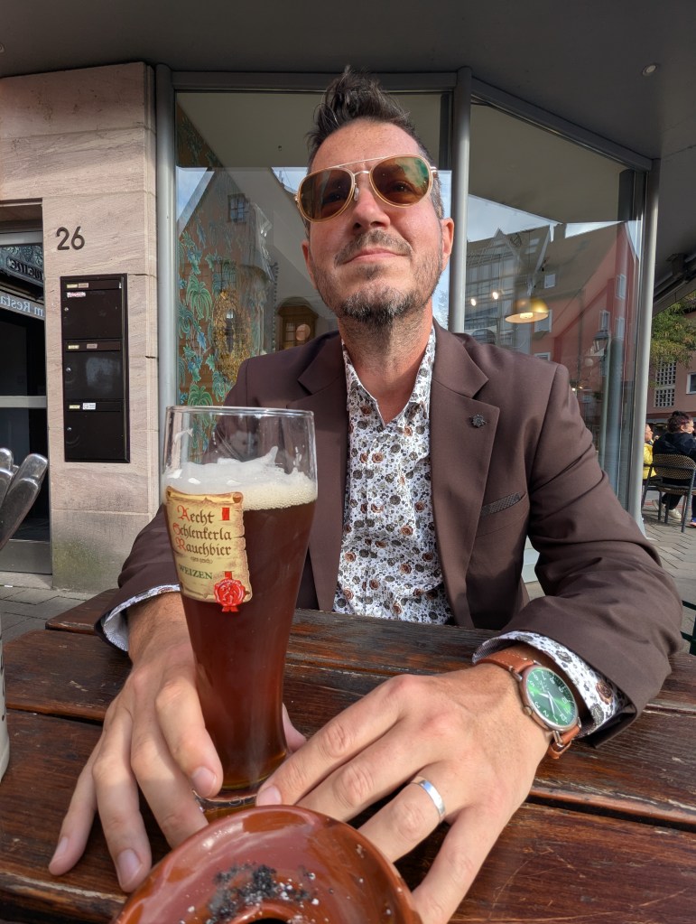 A man wearing sunglasses and a brown blazer sits at a table outdoors, smiling while holding a glass of dark beer. He has a floral shirt underneath the blazer and a watch on his wrist, with a small dish of ashes on the table.