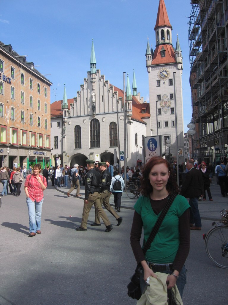 A young woman stands in the foreground wearing a green shirt, smiling at the camera. Behind her is a busy square featuring historical buildings with green-topped spires and a clock tower. People are walking and milling about, creating a lively atmosphere.