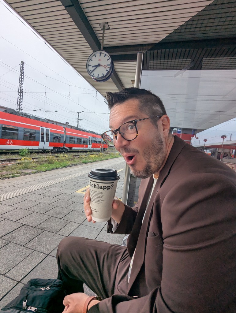 A man in a suit sits at a train station, holding a coffee cup with a surprised expression. A train is visible in the background, and a clock is mounted on the station roof.