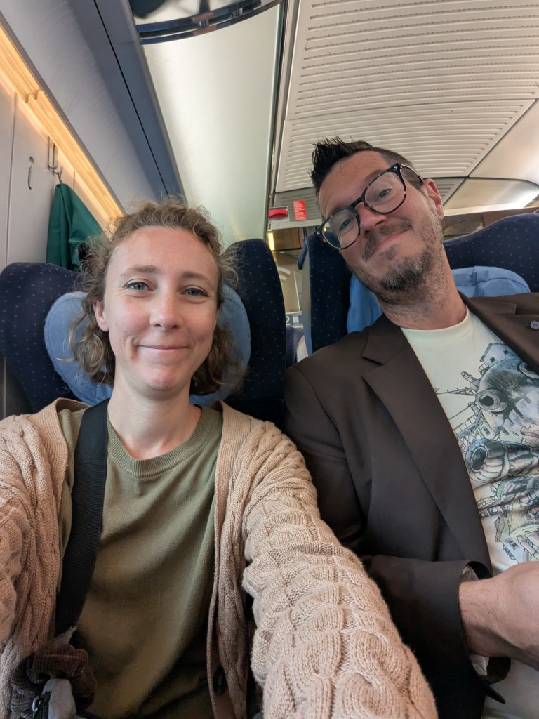 A man and a woman smiling together while seated in a train, with a cozy interior visible in the background.
