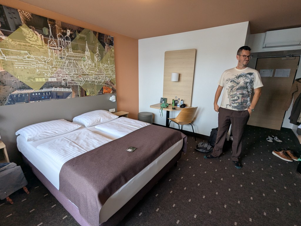 A hotel room featuring a large bed with white linens and a brown throw blanket, a man standing nearby in casual attire, and a modern aesthetic with a mural on the wall and minimalist furniture.
