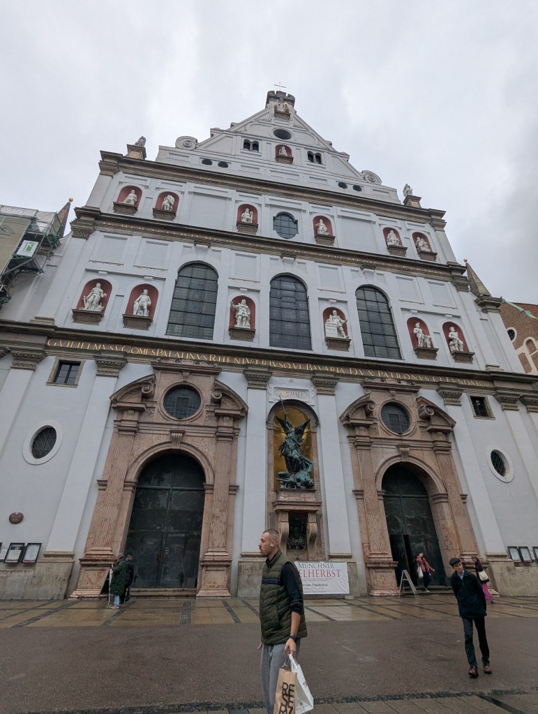 A large historical building with a decorative façade featuring statues and large arched doors. In front of the building, people are walking, and a banner is displayed. The sky is overcast.