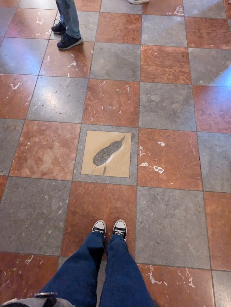 View of a tiled floor with a decorative inset design, showing feet of a person wearing sneakers and jeans.