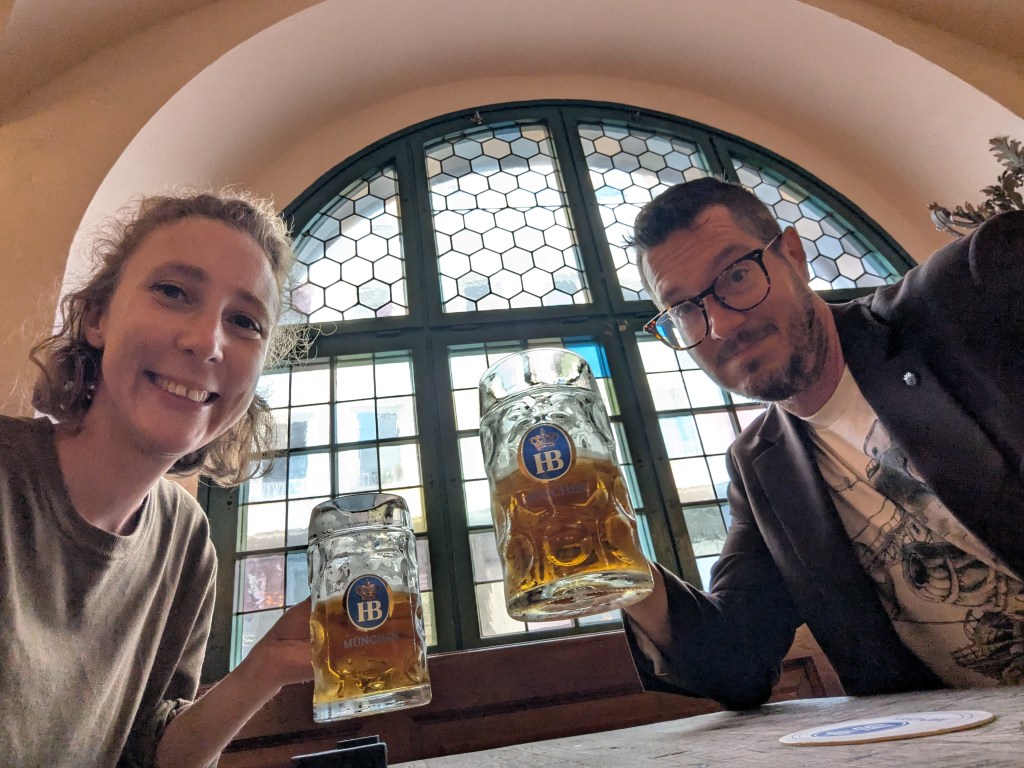 Two people smiling and holding large beer steins in a cozy establishment, with a stained glass window in the background.