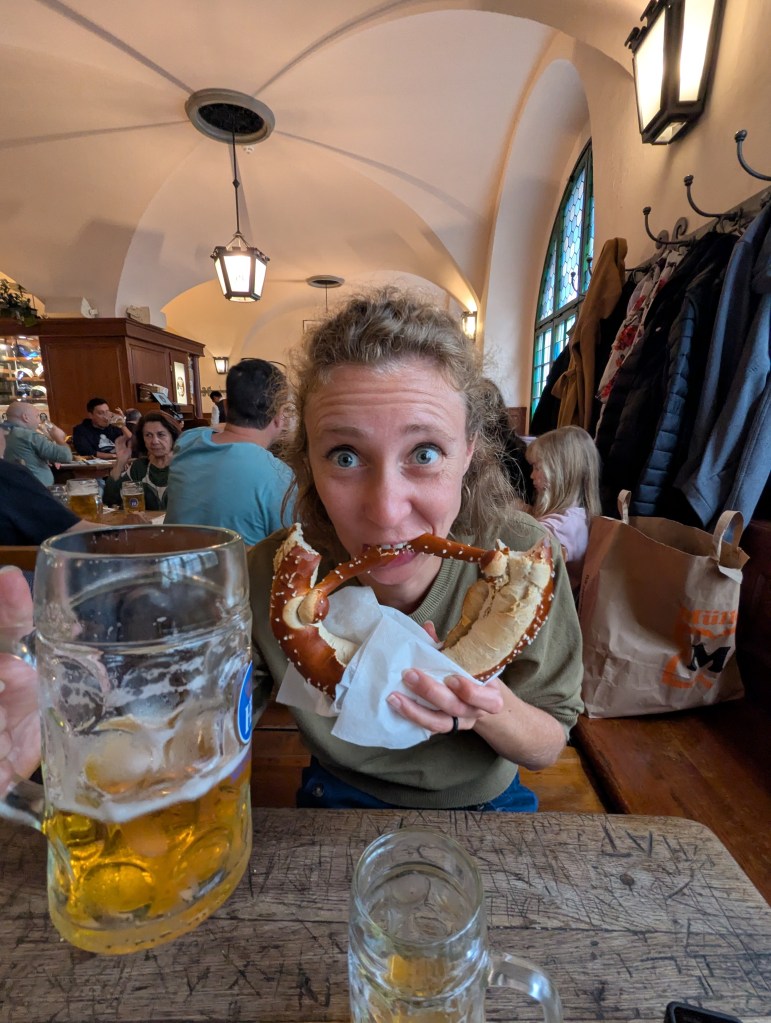 A smiling woman holding a large pretzel in front of her face, with a beer mug in the foreground. The setting is a lively pub with people dining in the background.