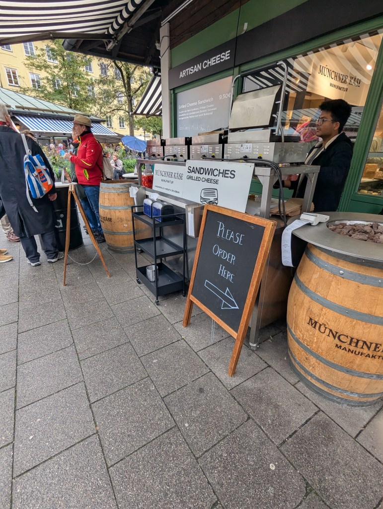 A food stall at an outdoor market featuring a sign for 'Artisan Cheese' and a menu for 'Grilled Cheese Sandwiches.' A chalkboard sign nearby indicates 'Please Order Here,' with people waiting in line.