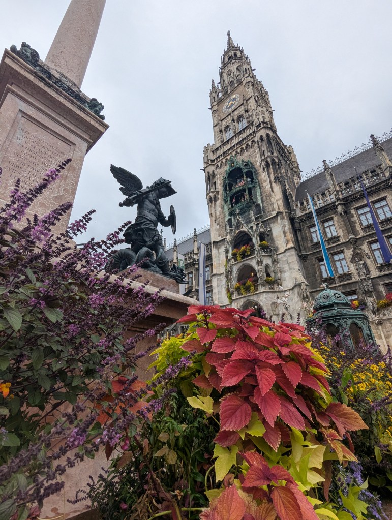 A low-angle view of a historic building with a tall clock tower, surrounded by vibrant flowers and a statue of an angel holding a shield, set against a cloudy sky.