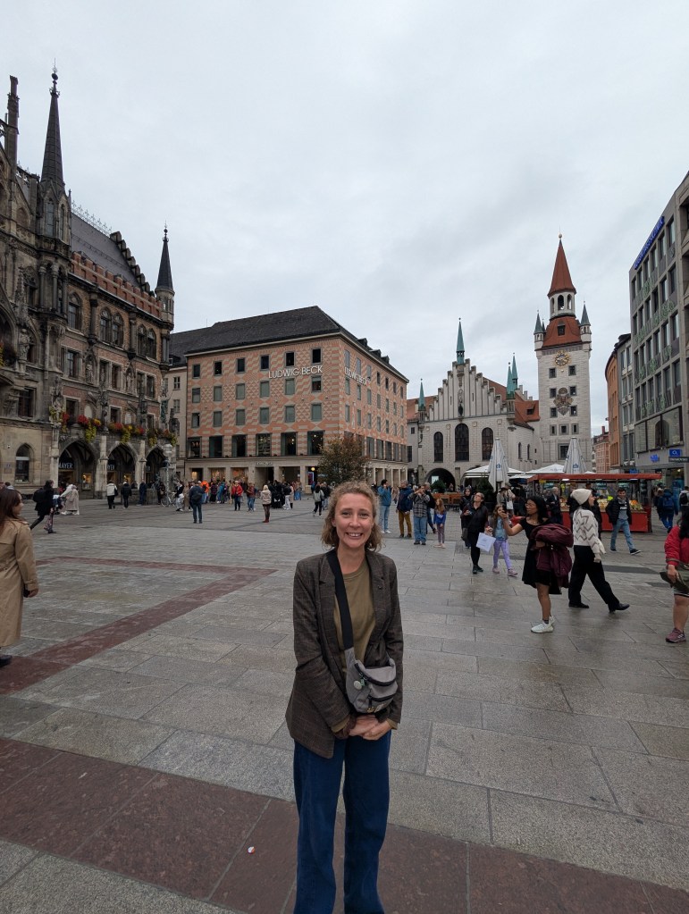 A woman standing in a bustling square with historic buildings, including a church and shops, against a cloudy sky.