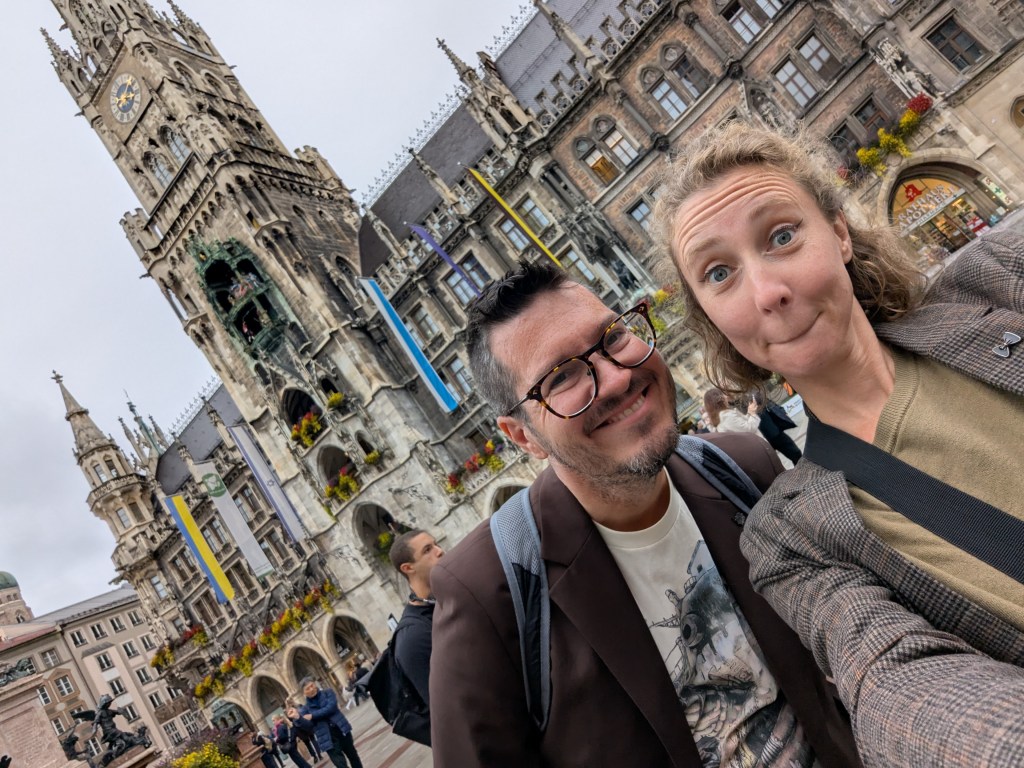 Two smiling individuals posing for a selfie in front of a historic building with ornate architecture and flower decorations, located in an urban setting.