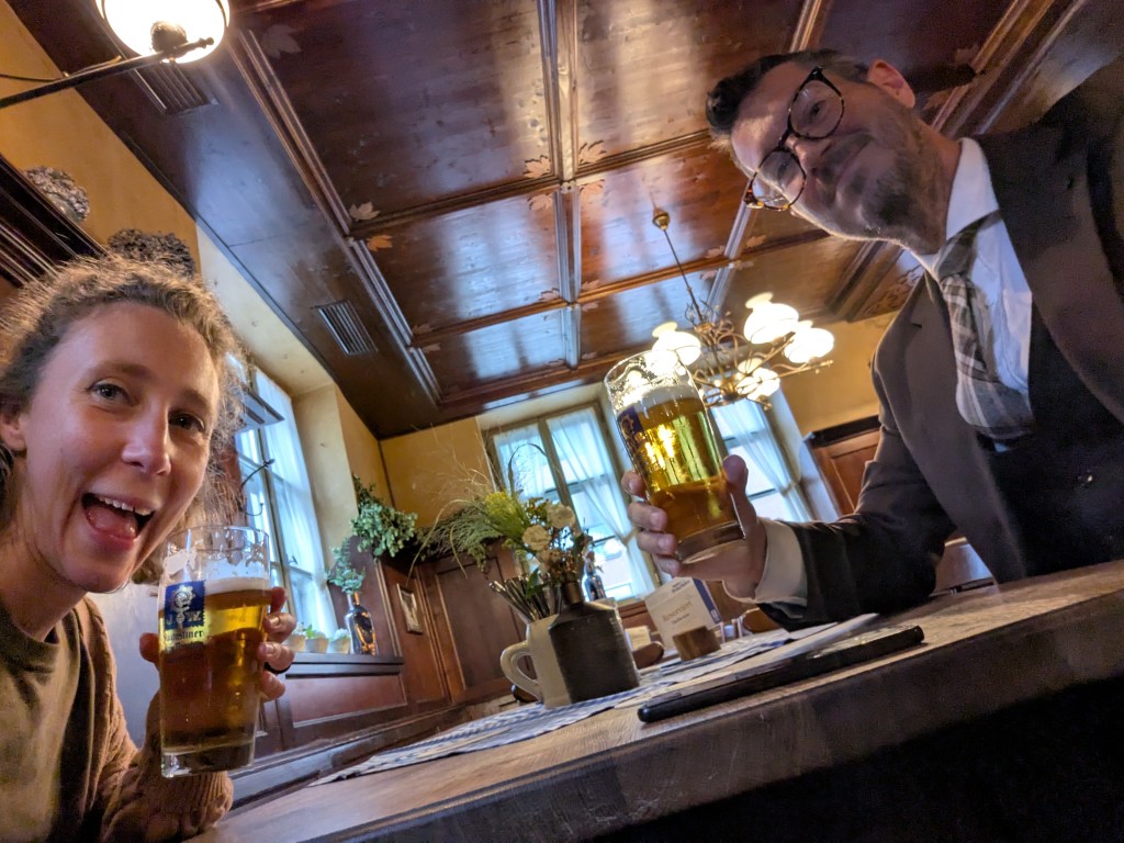 A man and a woman sitting at a table in a cozy pub, both holding large glasses of beer and smiling at the camera. The interior features wooden beams and warm lighting.