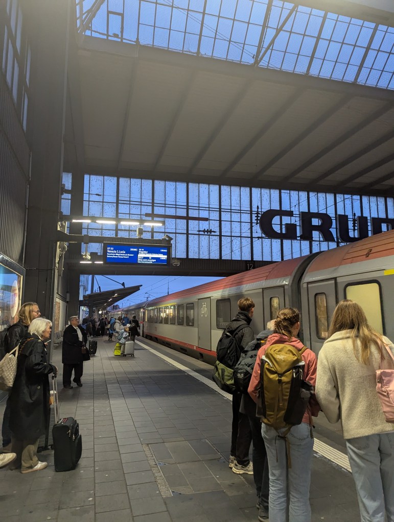 A busy train station platform with passengers waiting for a train. A digital display board shows train information, and a train is partially visible on the platform. The sky is darkening, indicating evening time.