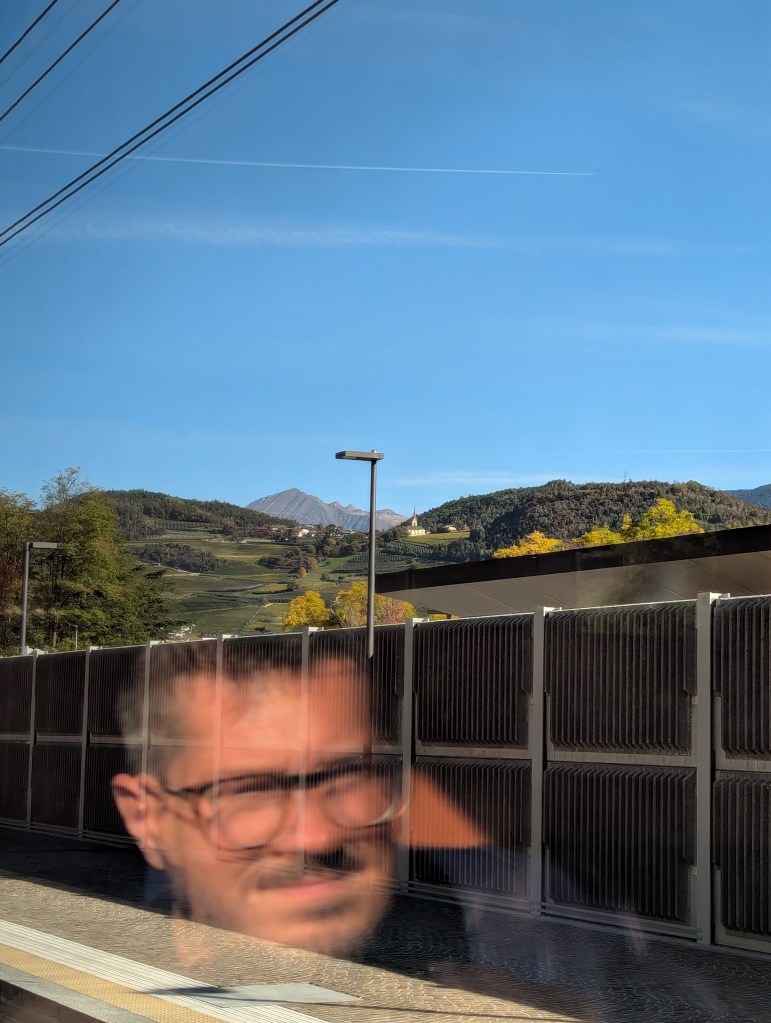 A reflection of a man with glasses and a mustache in a train window, with a mountainous landscape and blue sky in the background.