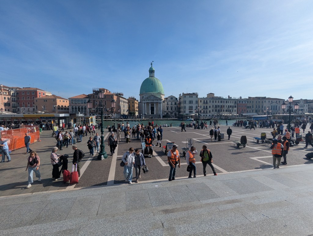 A busy piazza in Venice with many tourists and locals walking around, the green-domed church visible in the background. Some individuals are carrying luggage while others are engaged in conversations. There are several people in orange vests, indicating they might be part of the service or security team.