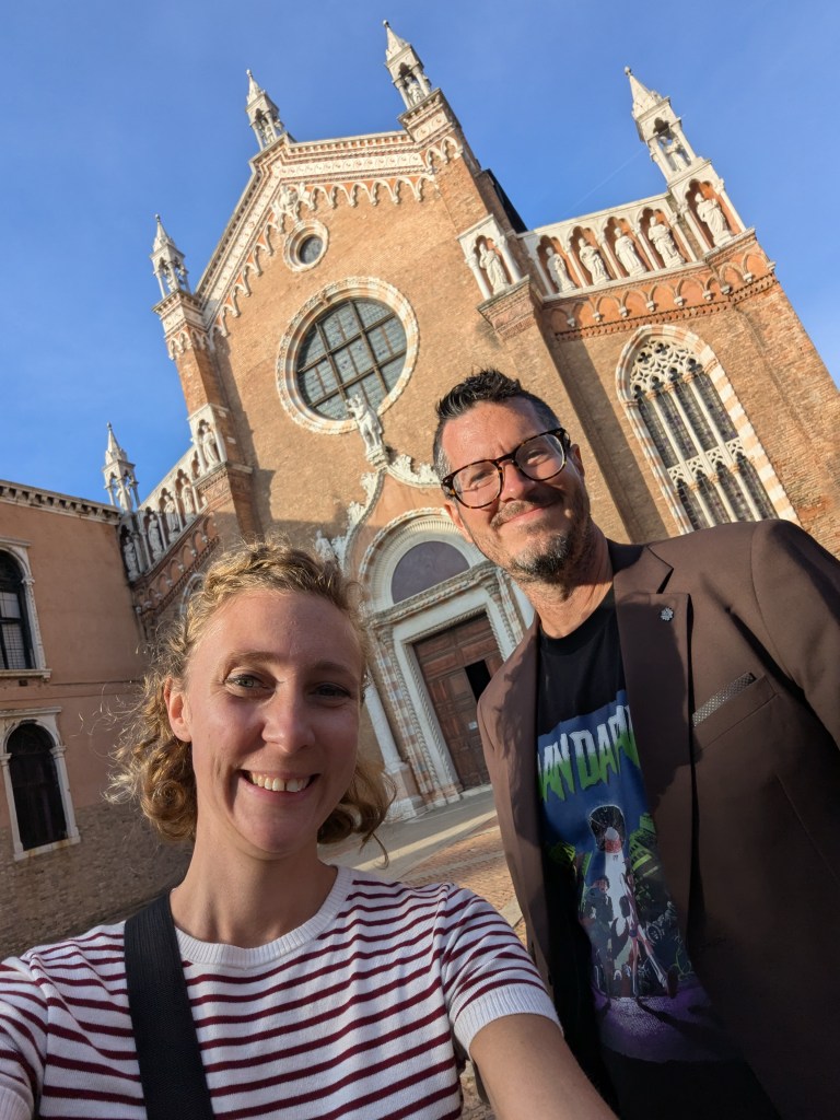 Selfie of a woman and a man in front of a historic brick building with ornate architecture and a large window.