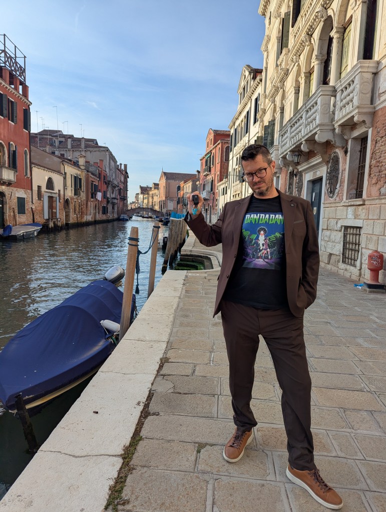 A man in a brown suit and graphic t-shirt stands by a canal in Venice, Italy, with colorful buildings and a blue boat in the background.