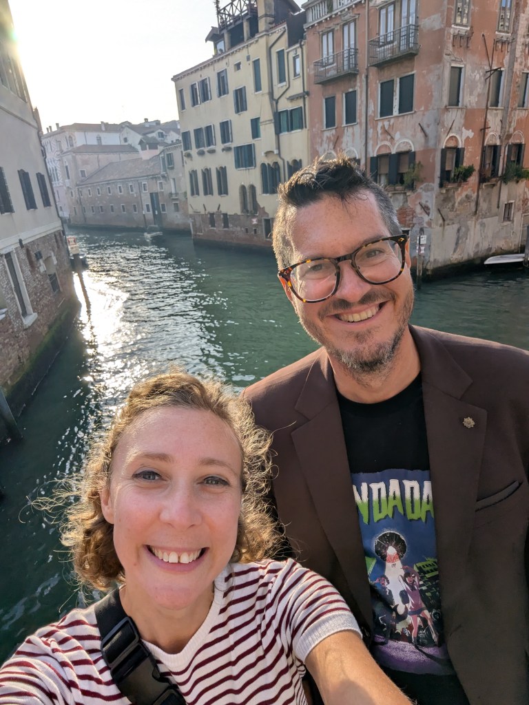 A cheerful selfie of a woman with curly hair wearing a striped shirt and a man with glasses in a brown jacket, against a scenic canal with buildings in Venice.