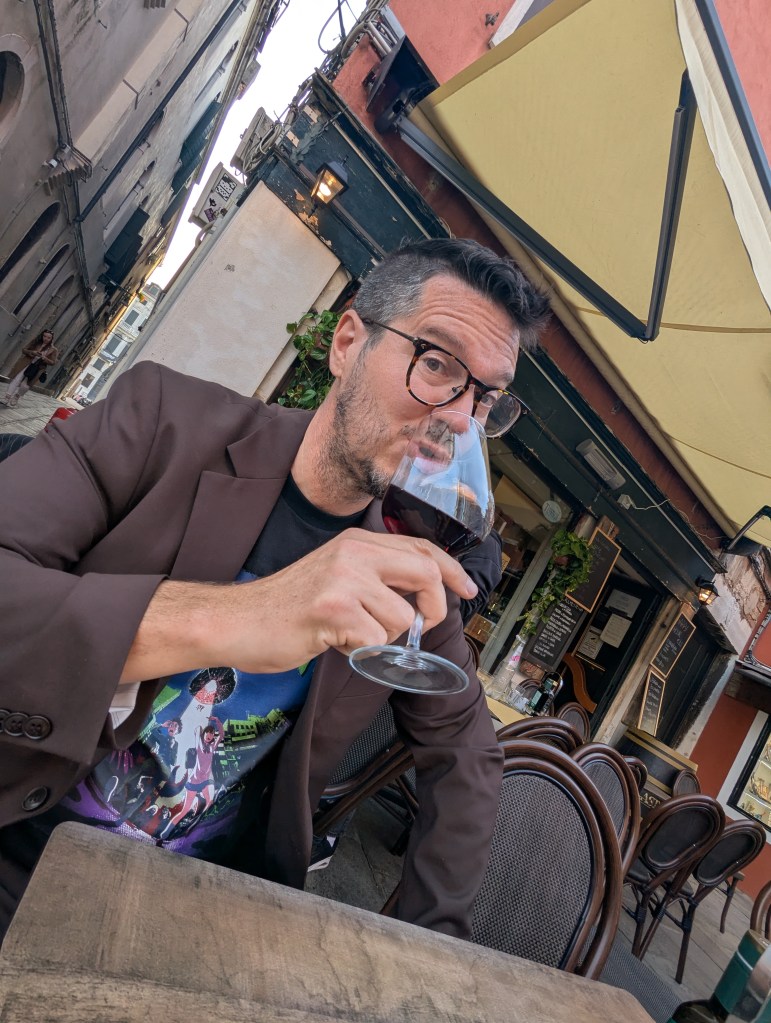 Person sitting at a table outdoors, enjoying a glass of red wine, wearing glasses and a black graphic t-shirt under a brown blazer, with a street scene in the background.