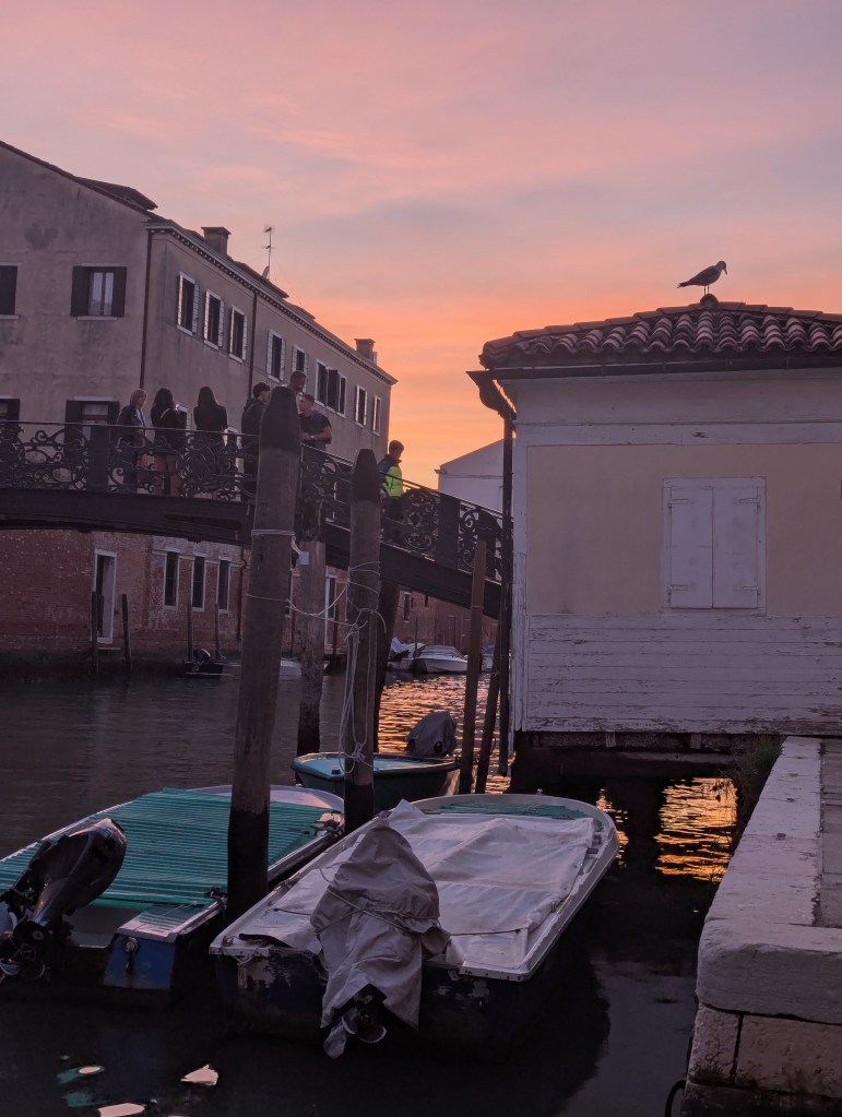 A picturesque scene at sunset in Venice, featuring a canal with moored boats, a bridge with people crossing, and a seagull perched on a nearby rooftop.