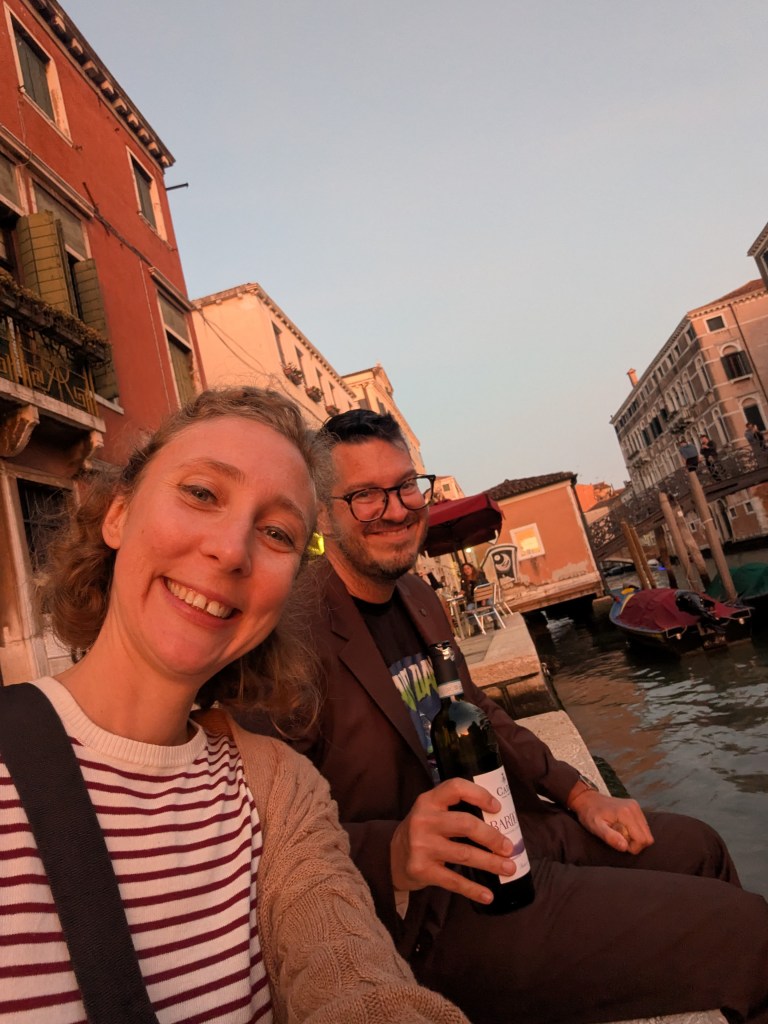 A smiling woman and man sitting by a canal in Venice during sunset, with buildings in the background. The man holds a bottle of wine.