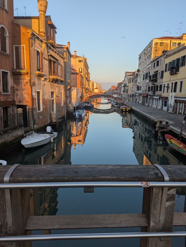 A serene canal scene in Venice at sunrise, featuring colorful buildings reflecting on the water's surface, with boats docked along the sides.