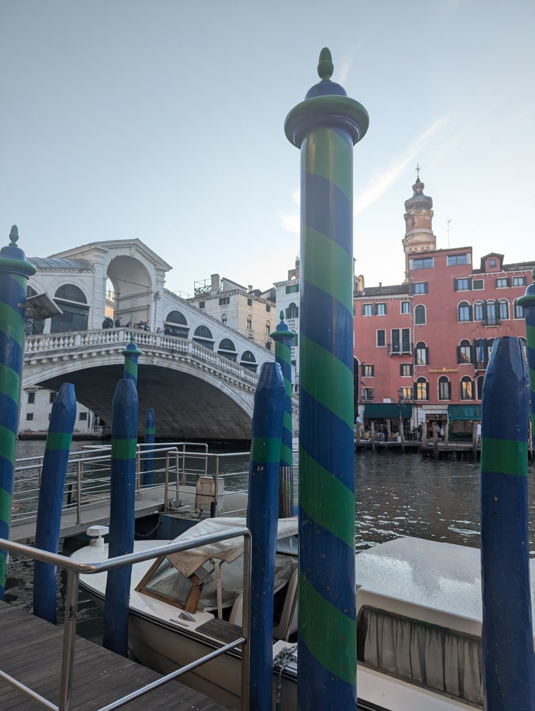 View of colorful mooring pylons along the canal in Venice, with the iconic Rialto Bridge in the background and historic buildings lining the waterfront.