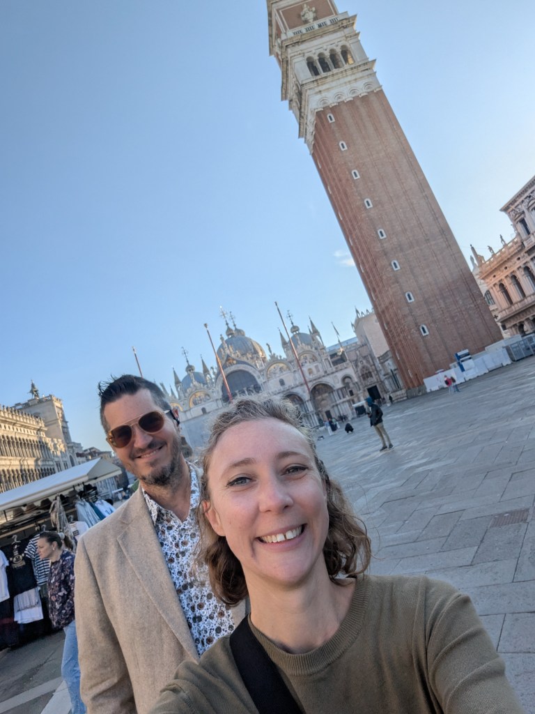 A selfie of two people in front of St. Mark's Campanile in Venice, Italy, with a clear blue sky in the background.