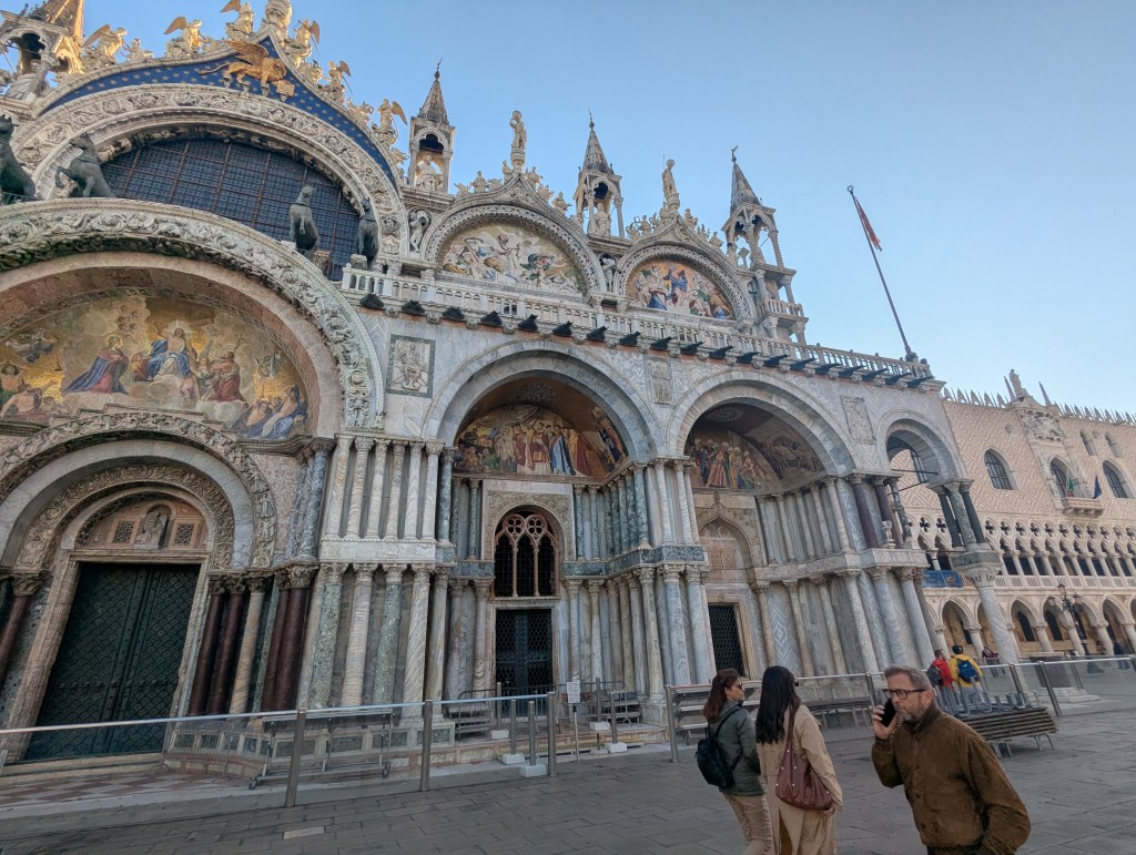 Exterior view of St. Mark's Basilica in Venice, showcasing its intricate architectural details and decorative mosaics, with pedestrians in the foreground.