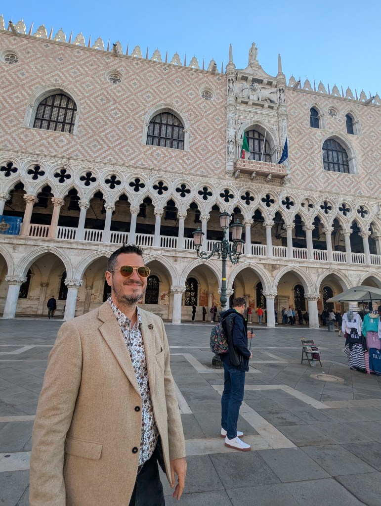 A man wearing sunglasses and a light-colored blazer stands in front of the ornate exterior of a historical building with intricate architectural details, featuring arched columns and decorative patterns. Other people can be seen in the background, and merchandise is displayed nearby.