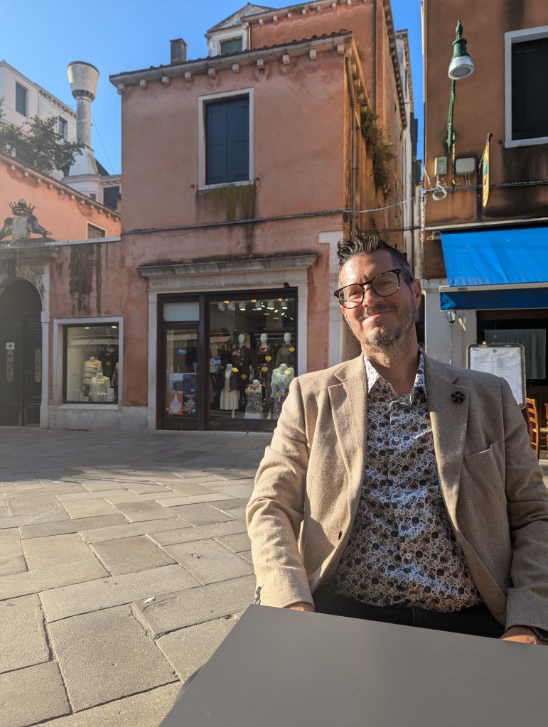 A man with glasses wearing a patterned shirt and blazer sits at a table in a sunny outdoor setting, with a backdrop of colorful buildings and shop windows.