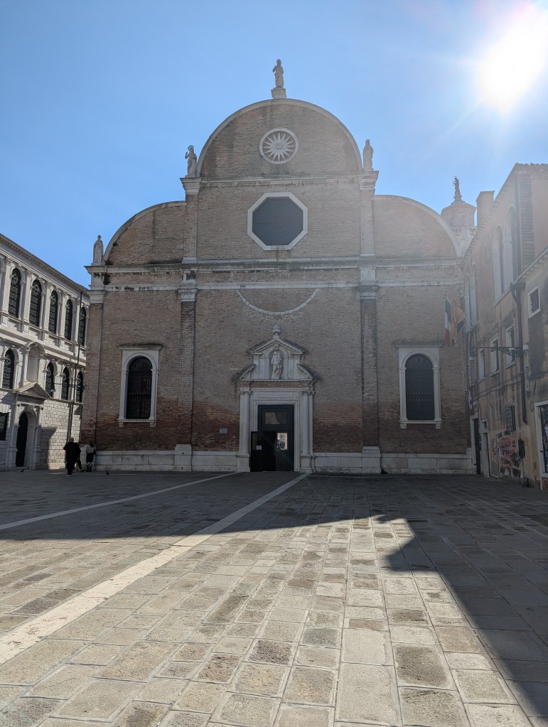 Exterior view of a historic church with a large circular window and statues on top, set against a clear blue sky. The foreground features a cobblestone square with a few people walking.