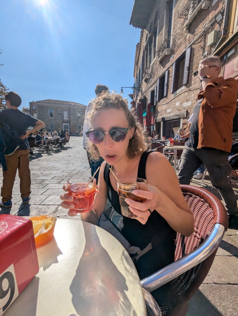 A woman sitting at a café table outdoors, holding two drinks, one red and one dark, with a sunny urban backdrop and people in the background.