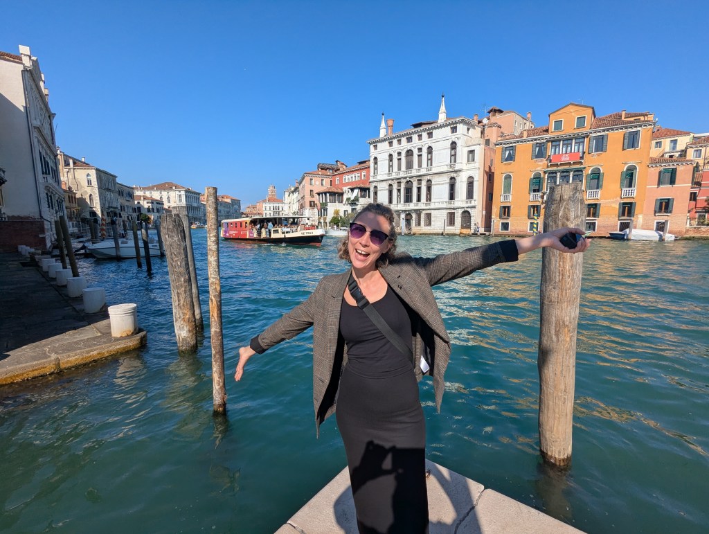 A woman in a gray jacket and sunglasses joyfully posing by a canal in Venice, Italy, with colorful buildings in the background and boats on the water.