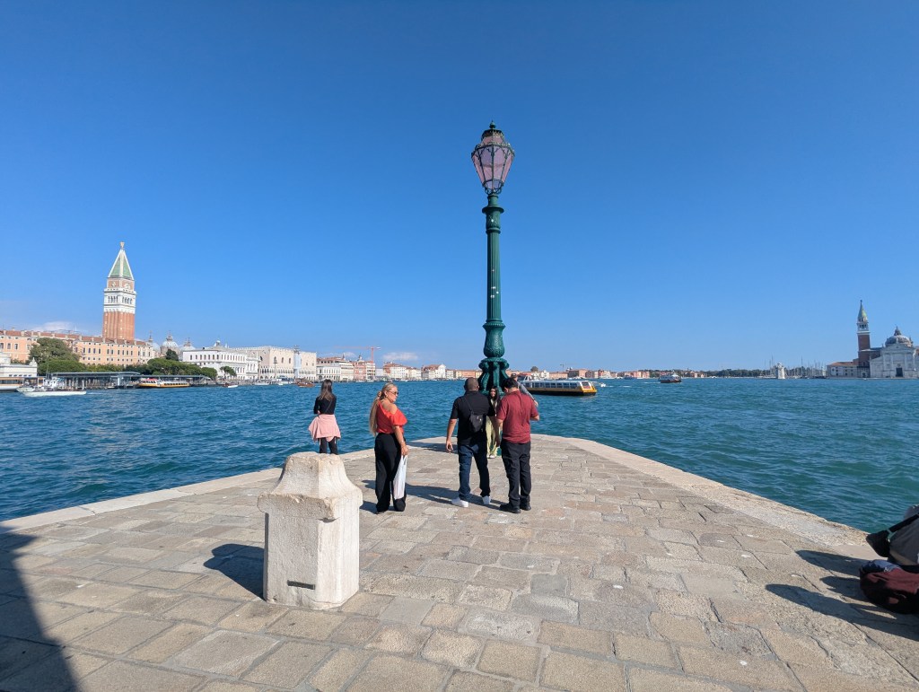 View of a waterfront pier in Venice, Italy, featuring a lamppost and several people admiring the scenery, with historic buildings and a clear blue sky in the background.